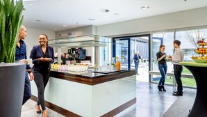 A modern hotel lobby with a central counter displaying food and beverages, a glass partition, and a view of the outdoors. - Novotel Köln City (Cologne)