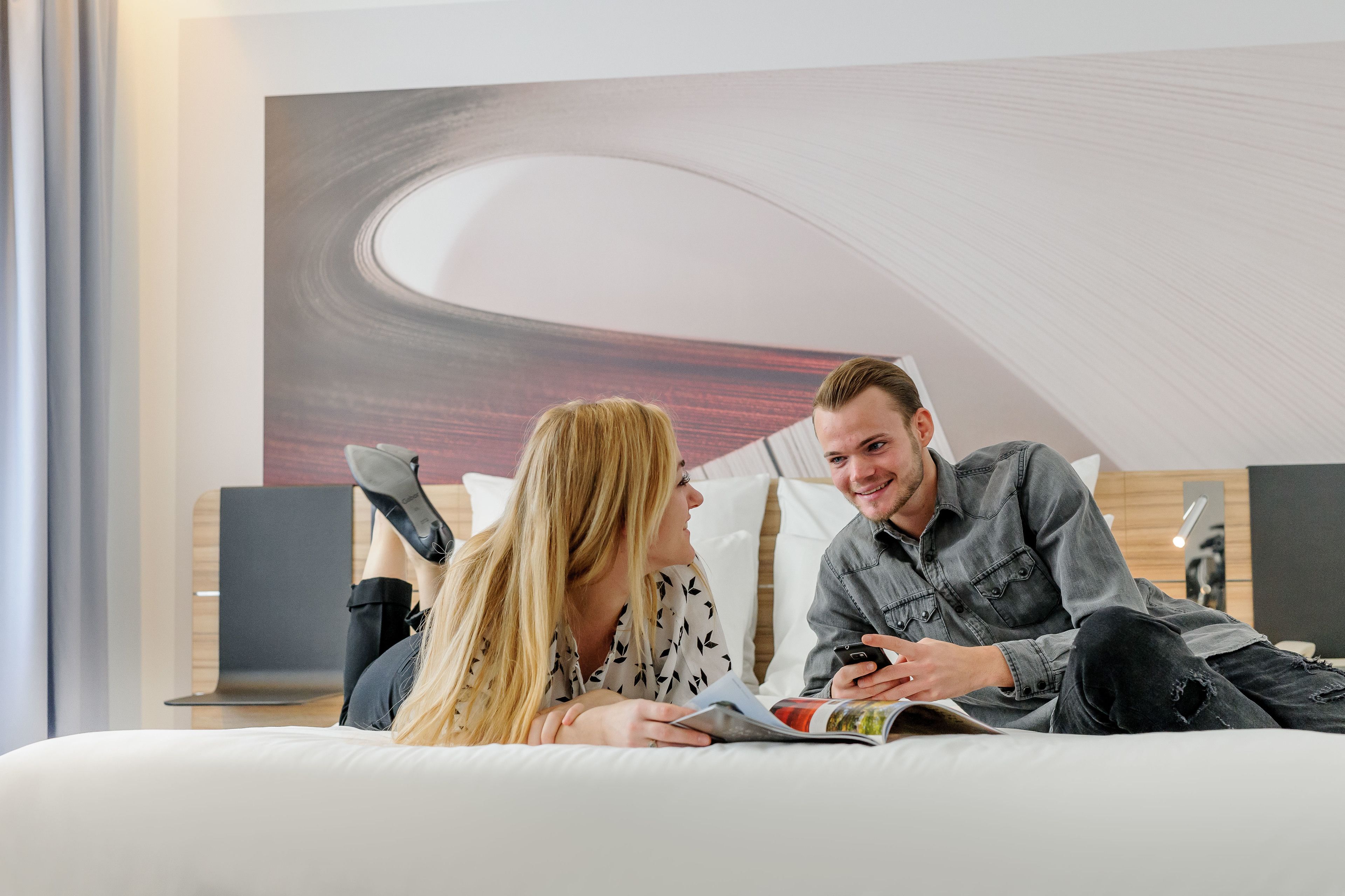 A modern room with a large abstract painting on the wall, a bed with white bedding, and a side table with a lamp.