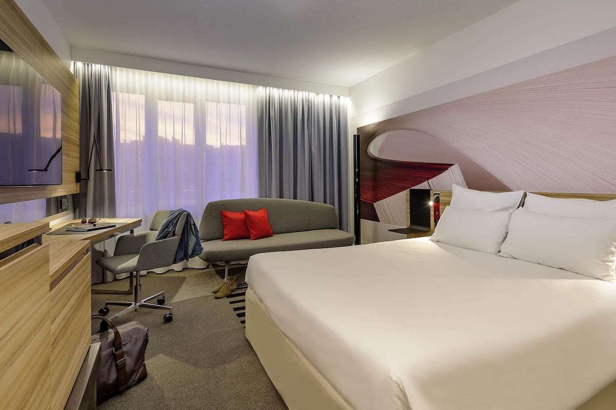 A hotel room with a bed, a desk, a chair, a TV, and a window with curtains.