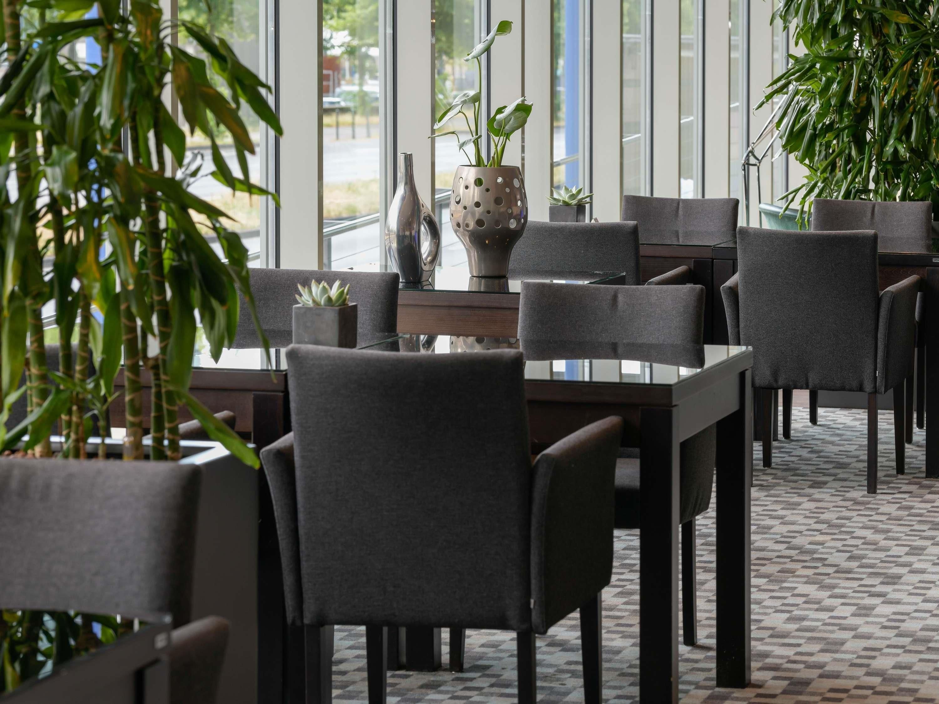 A restaurant with a modern interior, featuring dark-toned chairs, a glass table, and potted plants.