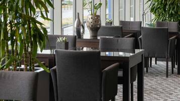A restaurant with a modern interior, featuring dark-toned chairs, a glass table, and potted plants.