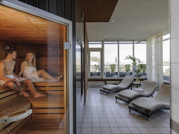 A sauna room with wooden benches and a glass door leading to a balcony with lounge chairs and a city view.