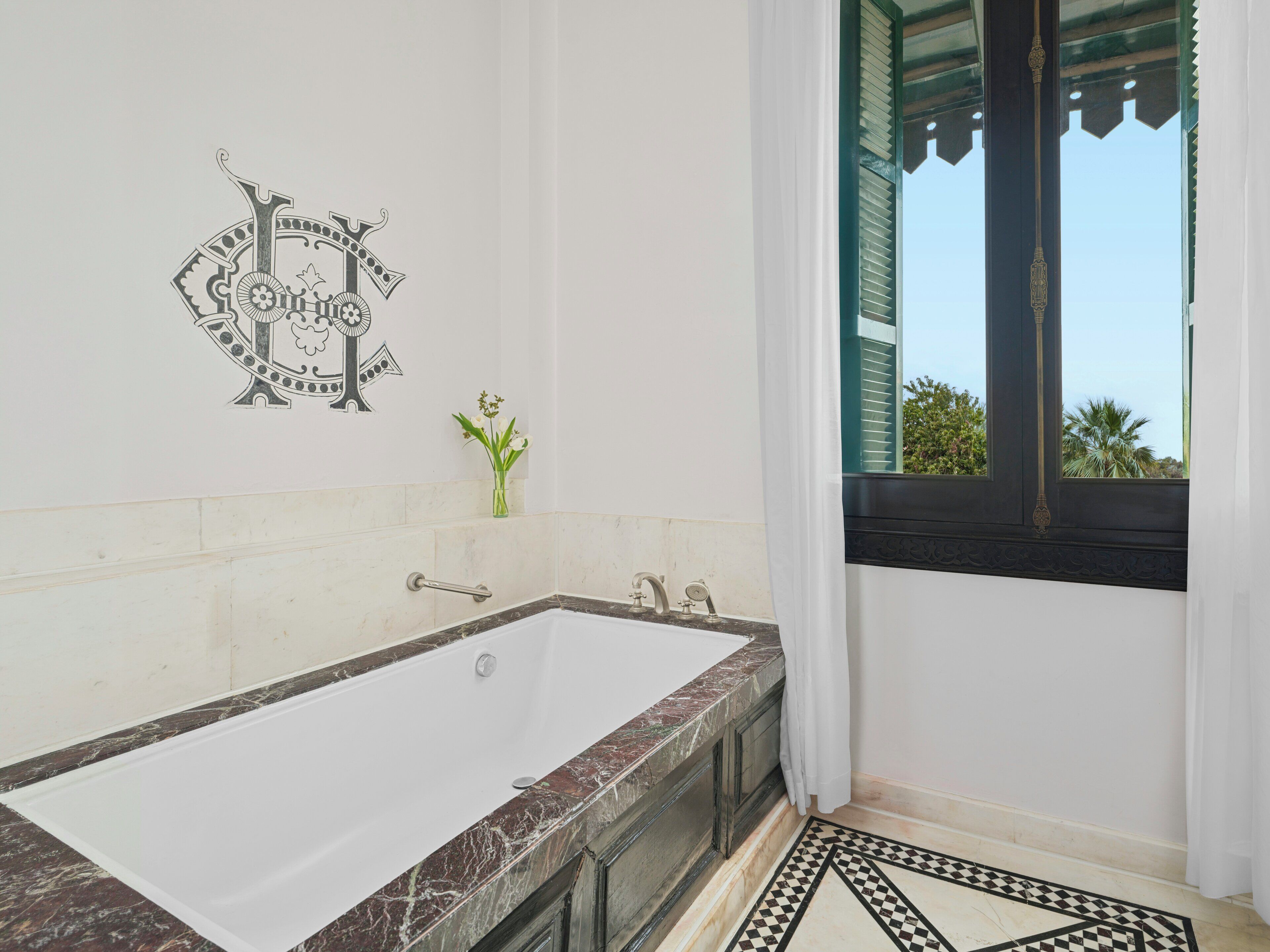 Un baño con una bañera grande, un jarrón pequeño con flores y una ventana con vistas a palmeras.