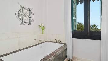 Un baño con una bañera grande, un jarrón pequeño con flores y una ventana con vistas a palmeras.