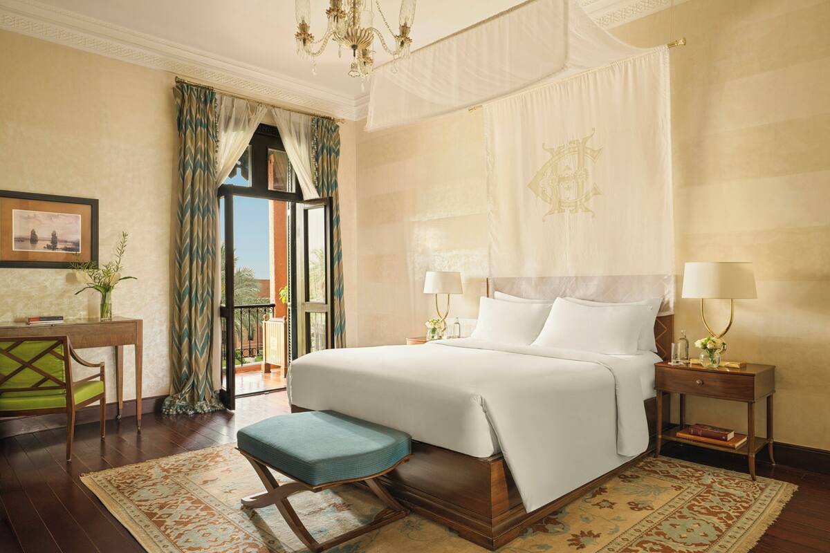 A hotel room with a large bed, bedside tables, a desk, a chair, and a balcony with curtains.