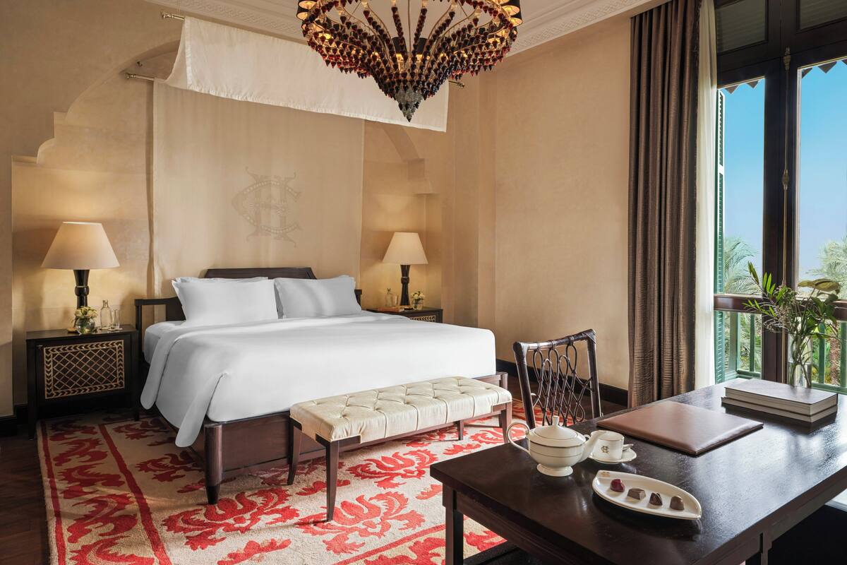 A hotel room with a bed, two bedside tables with lamps, a bench, a table with a teapot and cups, and a view of palm trees through a window.