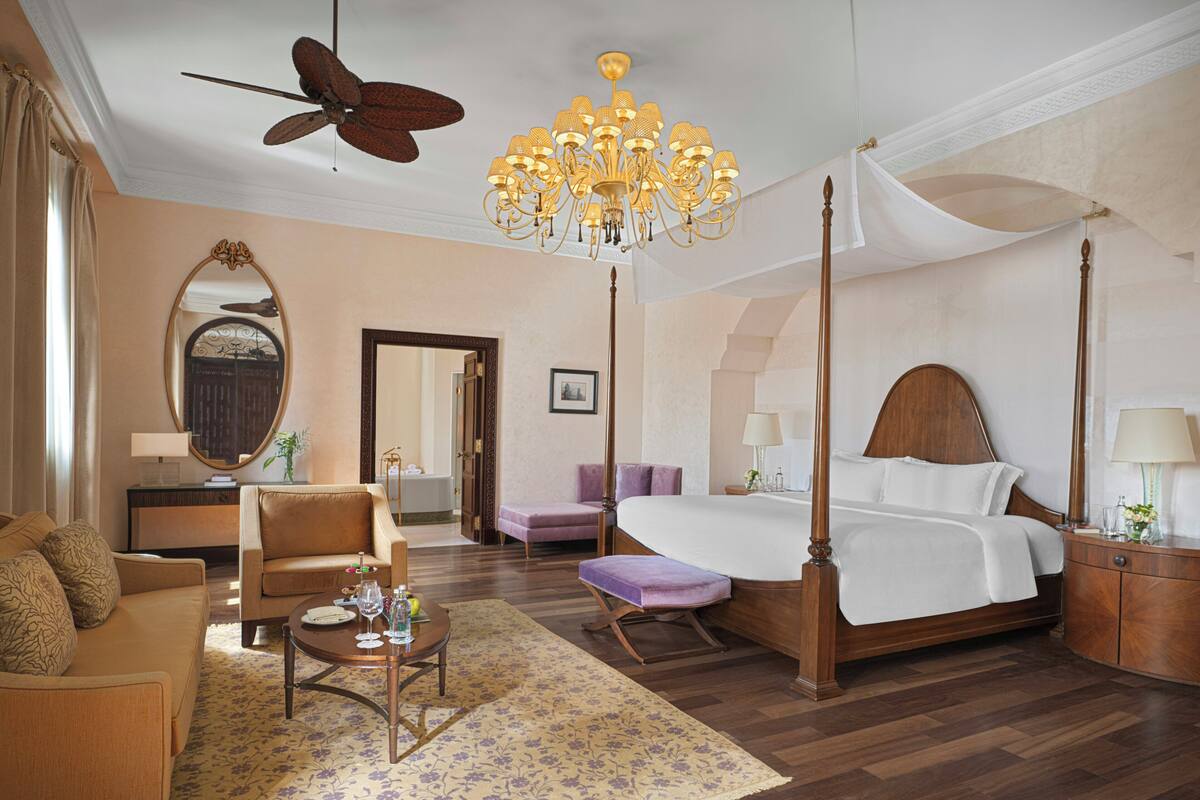 A four-poster bed, a sitting area with a sofa and armchair, a dining table with chairs, a chandelier, and a ceiling fan.