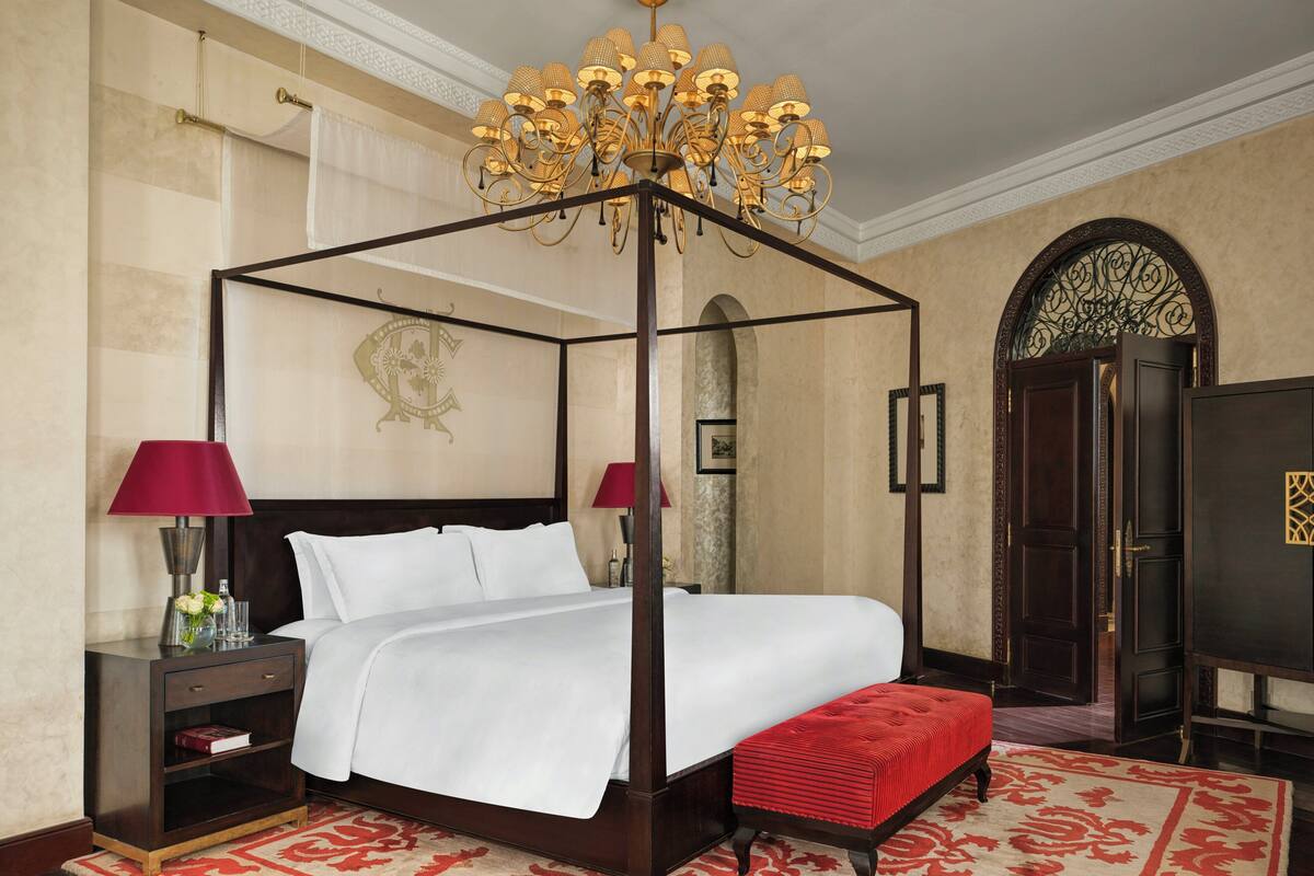 A four-poster bed with a red upholstered bench, a chandelier, a nightstand with a lamp, and a red rug.
