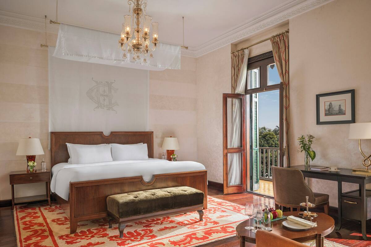 A hotel room with a large bed, a sitting area with a table, a desk, and a balcony with curtains.