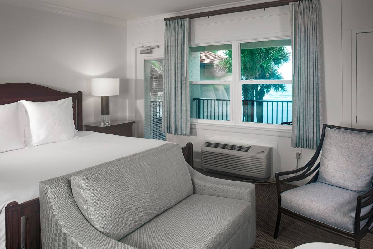 A hotel room with a bed, a sofa, a chair, a table with fruit, and a window with curtains.