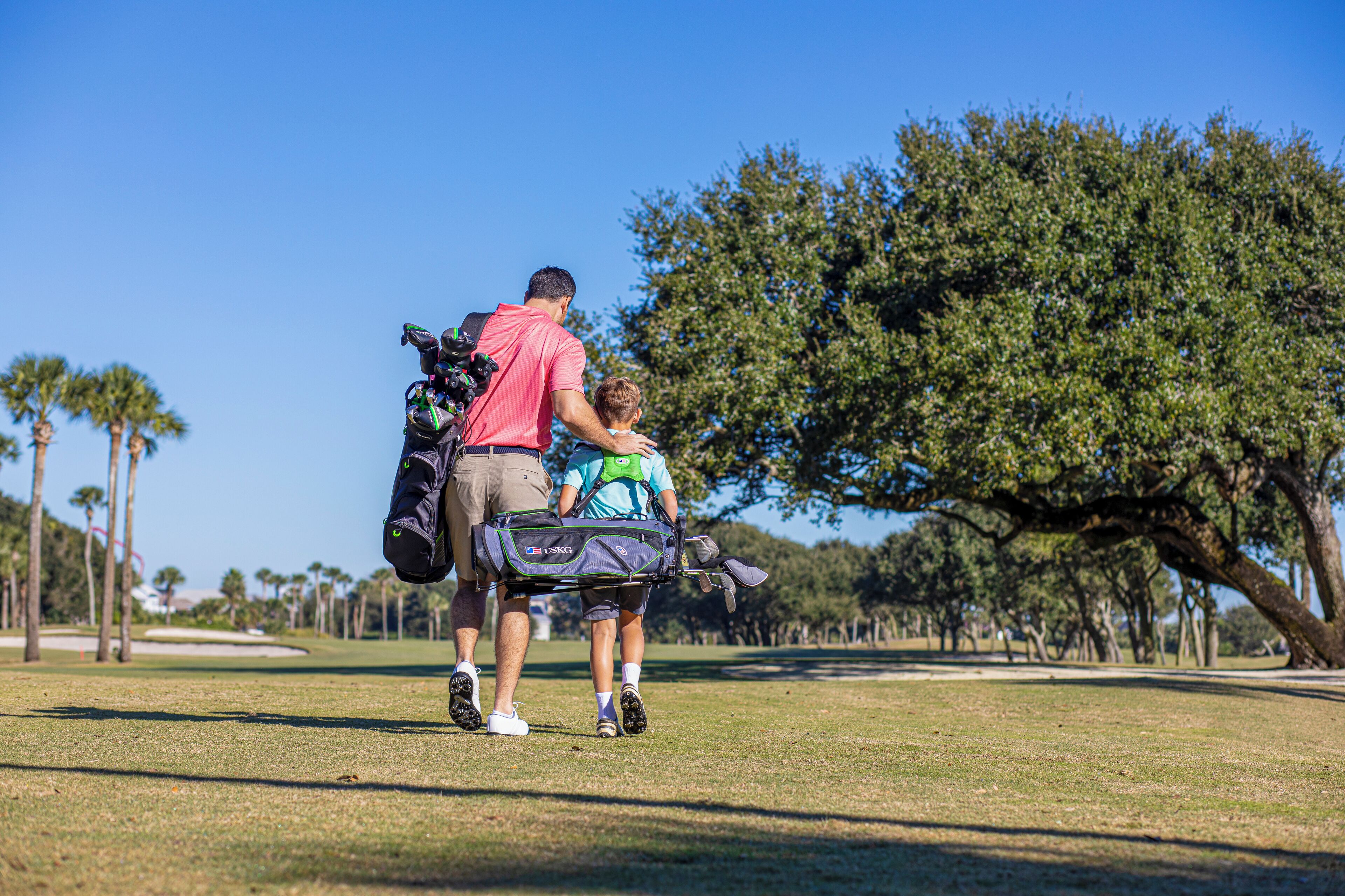 A man and a child walking on a golf course with a golf bag and a golf club.