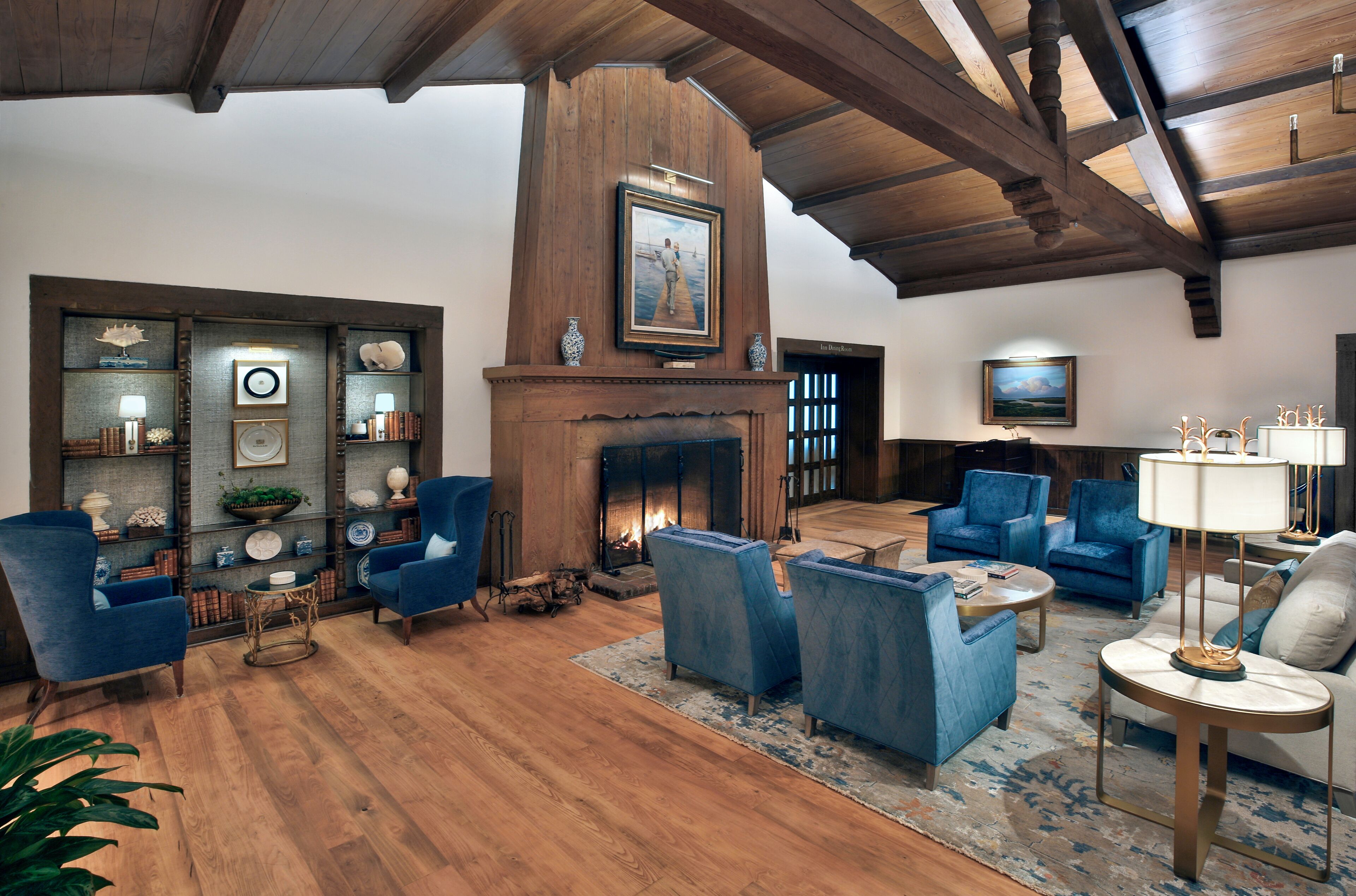 A spacious living room with a fireplace, blue armchairs, a coffee table, and a bookshelf with decorative items.