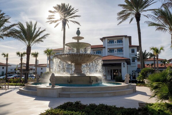 A multi-tiered fountain with water jets, surrounded by palm trees and a building with balconies.