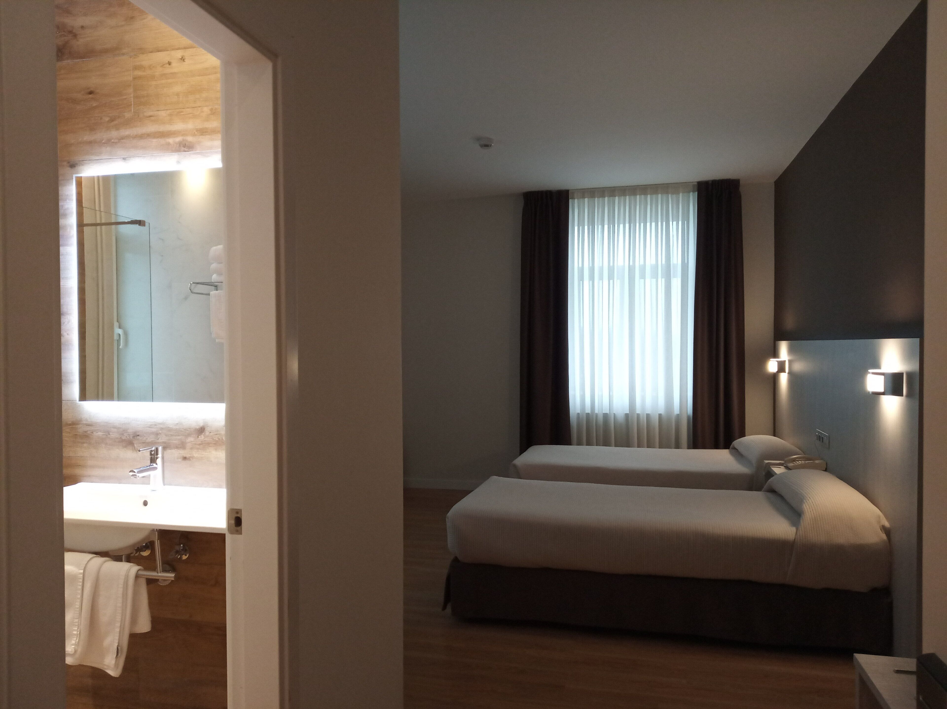 A hotel room with a wooden floor, a large mirror, a bathroom with a sink and towel rack, two beds, and a window with curtains.