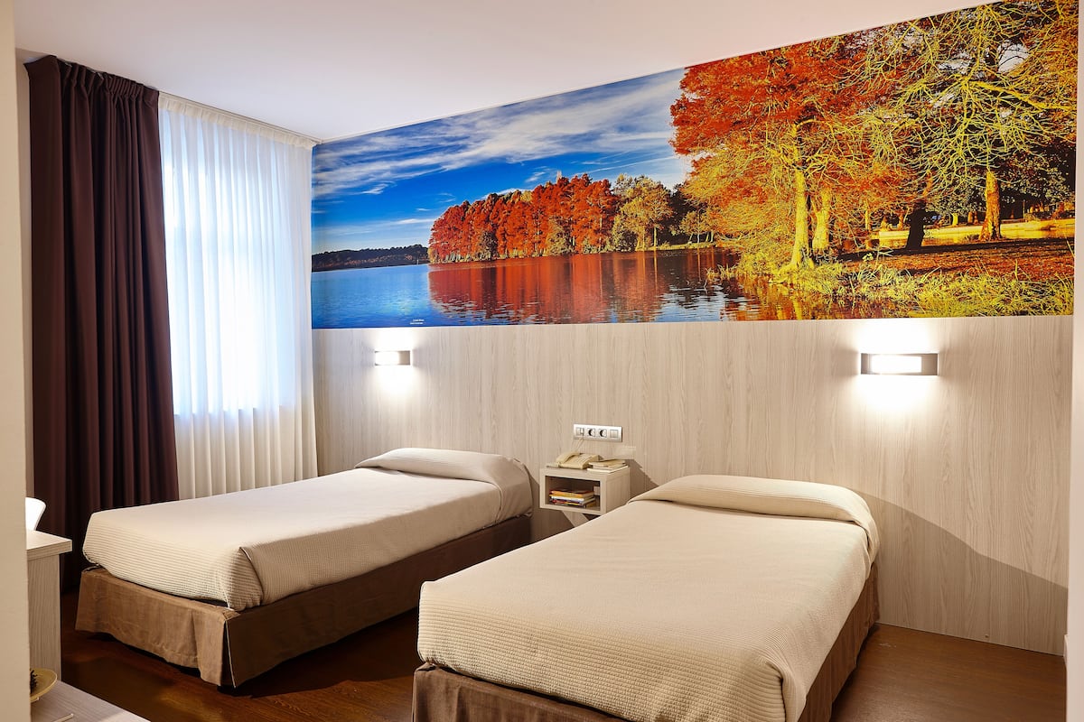Quarto de hotel com duas camas, uma mesa de cabeceira com telefone e livros, e um grande mural na parede retratando um lago e árvores de outono.