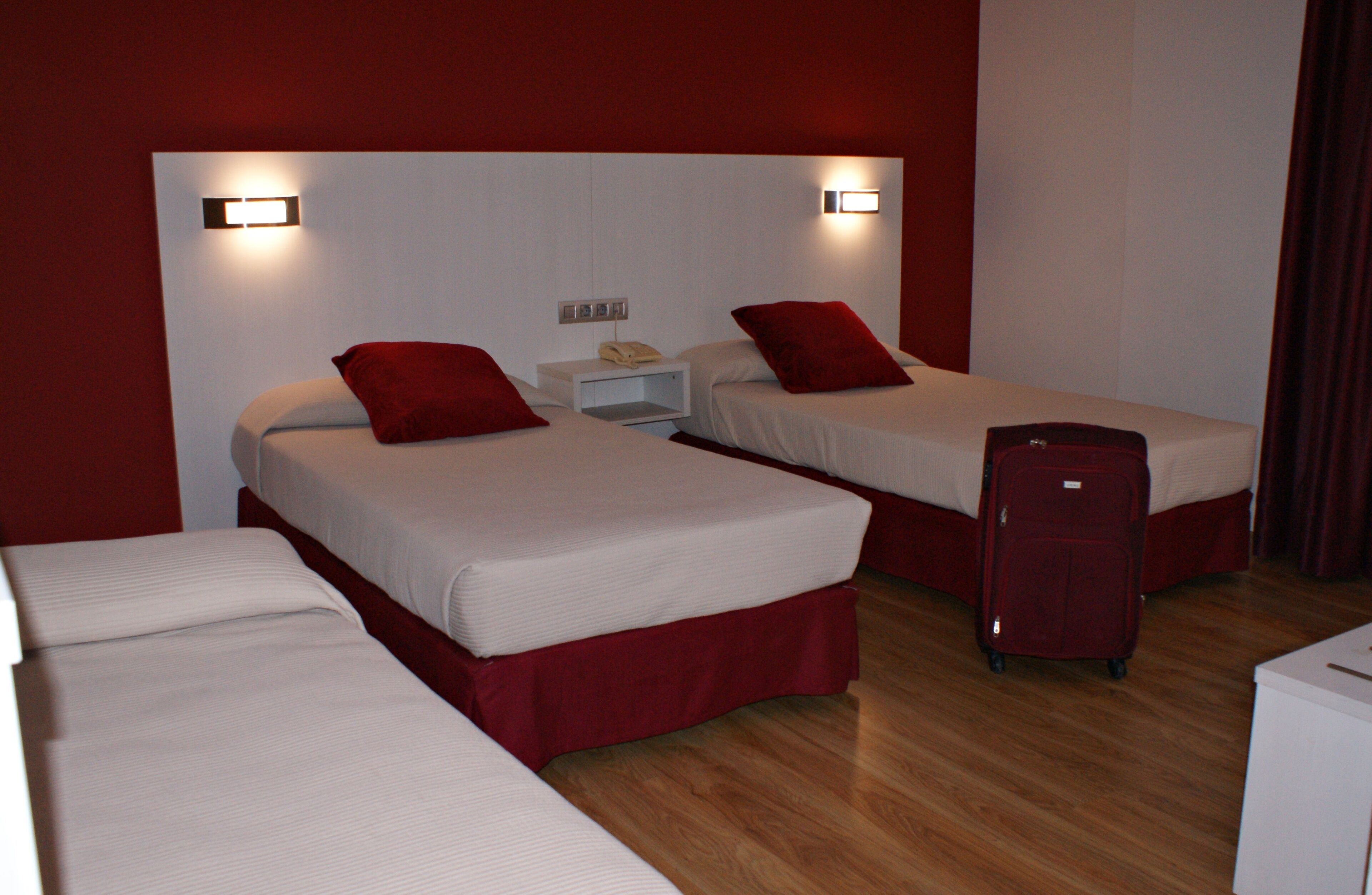 Une chambre d’hôtel avec deux lits simples, chacun agrémenté d’oreillers rouges et de draps blancs. On y trouve une table de chevet blanche avec un téléphone, et une valise rouge posée sur le sol.