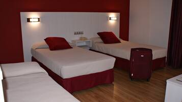 Une chambre d’hôtel avec deux lits simples, chacun agrémenté d’oreillers rouges et de draps blancs. On y trouve une table de chevet blanche avec un téléphone, et une valise rouge posée sur le sol.