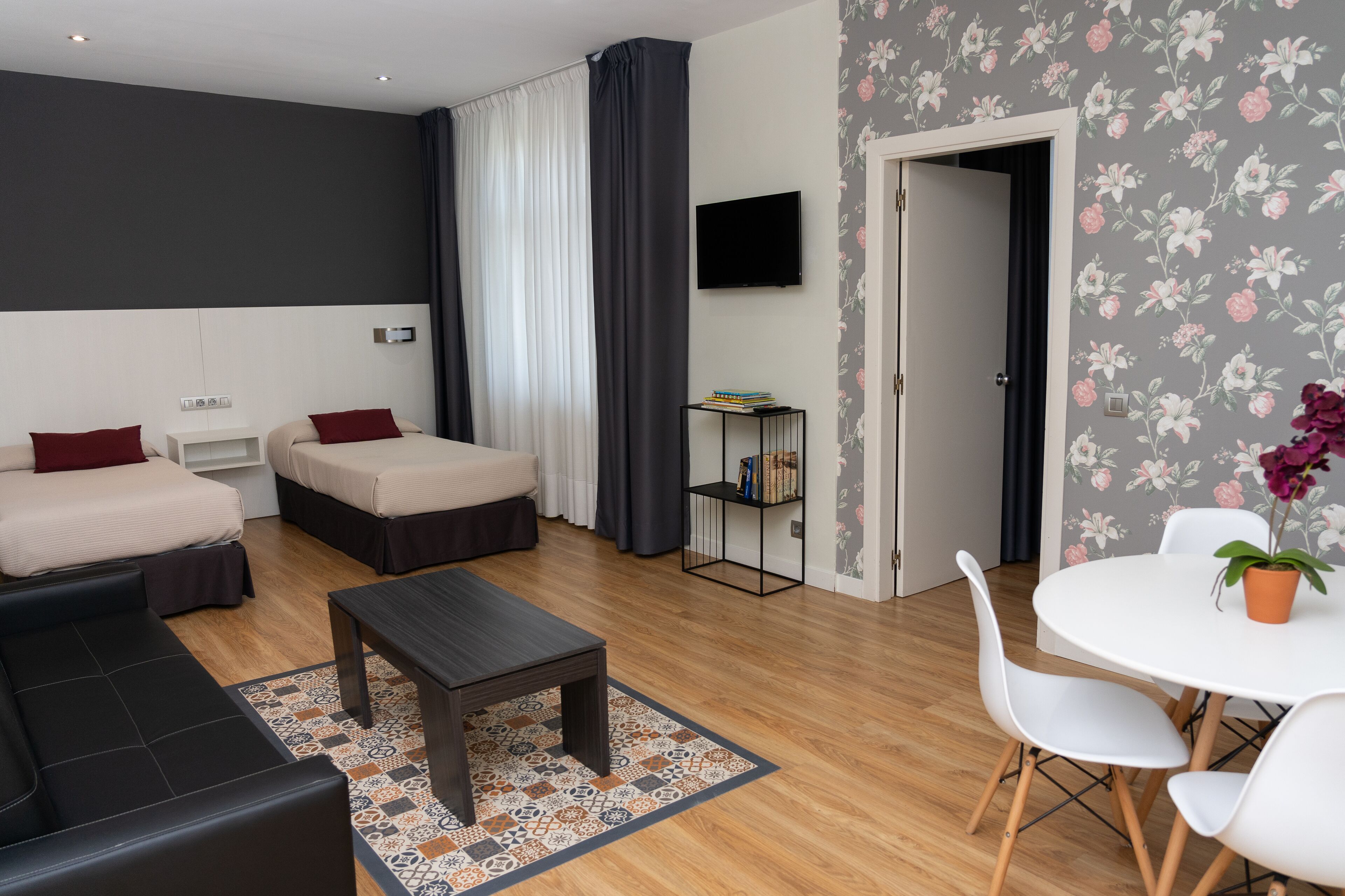 Una habitación de hotel moderna con sofá, dos camas individuales, una mesita, televisor y papel tapiz con motivos florales.