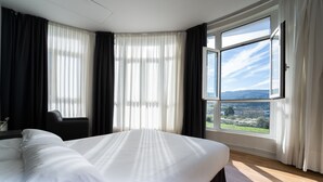 Una cama grande con ropa de cama blanca, cabecera oscura y vista a un paisaje a través de un ventanal amplio.
