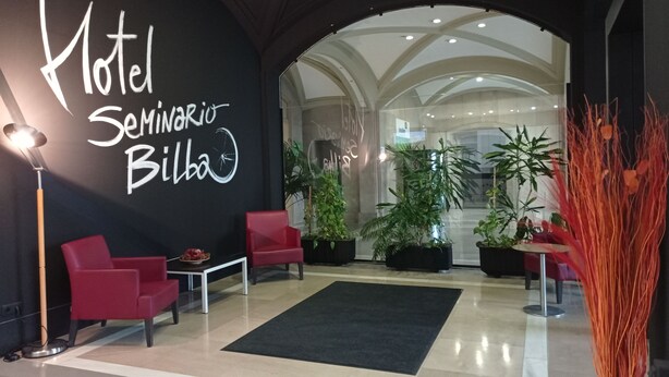 Un vestíbulo de hotel con un cartel grande que dice "Hotel Seminario Bilbao", sillas rojas, plantas en macetas y una alfombra negra.