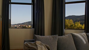 Habitación con vistas a un pueblo y colinas a través de amplios ventanales, un sofá con cojines grises y cortinas oscuras.