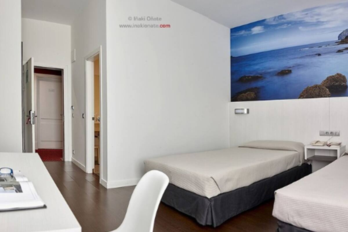 Quarto de hotel com duas camas de solteiro, uma escrivaninha com cadeira e uma janela grande com vista para a costa.