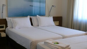 Quarto de hotel com duas camas, roupa de cama branca, pintura de um barco na água e uma janela com cortinas.