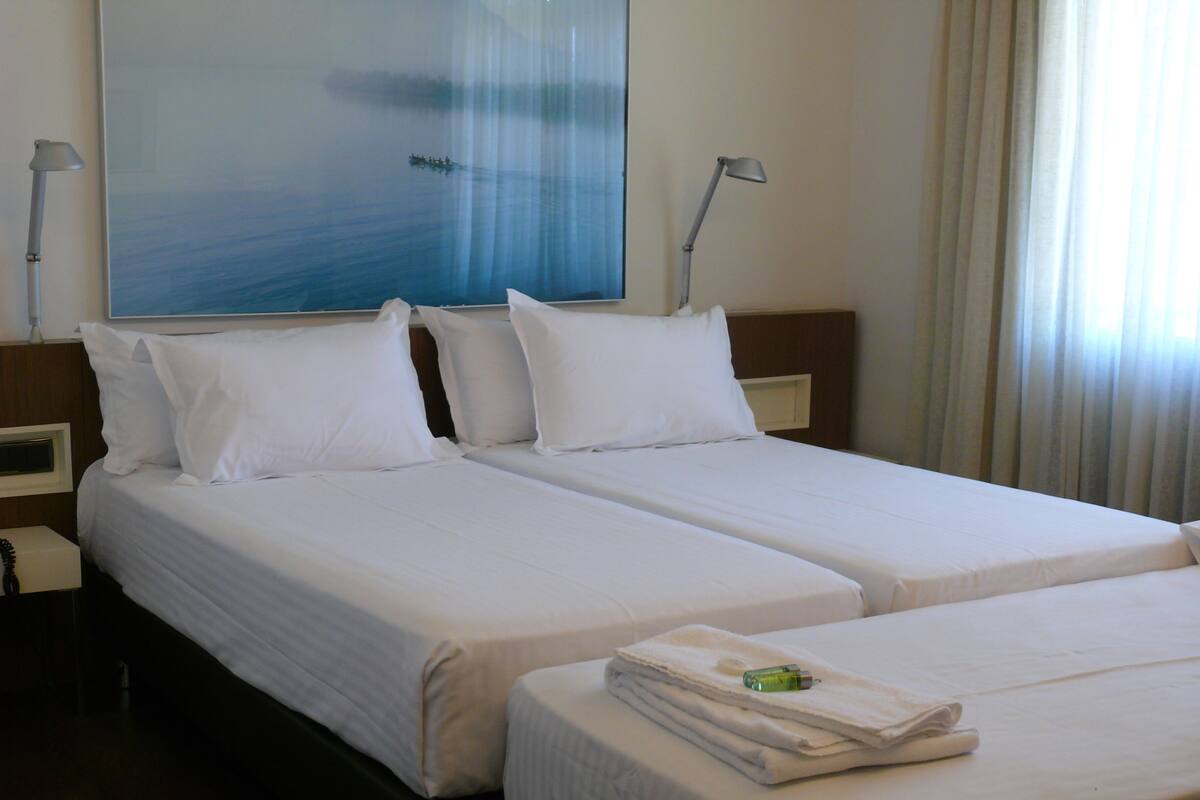 A hotel room with two beds, white bedding, a painting of a boat on water, and a window with curtains.