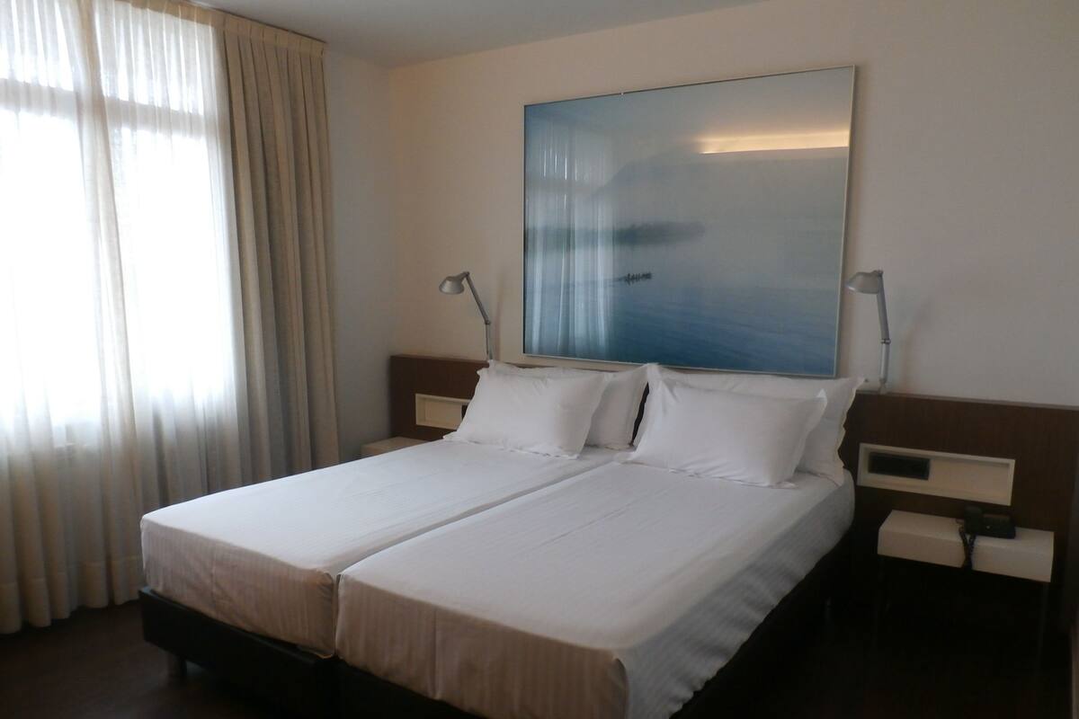 A double bed with white linens, a nightstand with a lamp, and a large abstract painting on the wall.