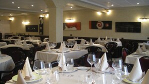 Um restaurante com mesas redondas preparadas para refeições, cada uma com toalha de mesa branca, guardanapos e copos de vinho.