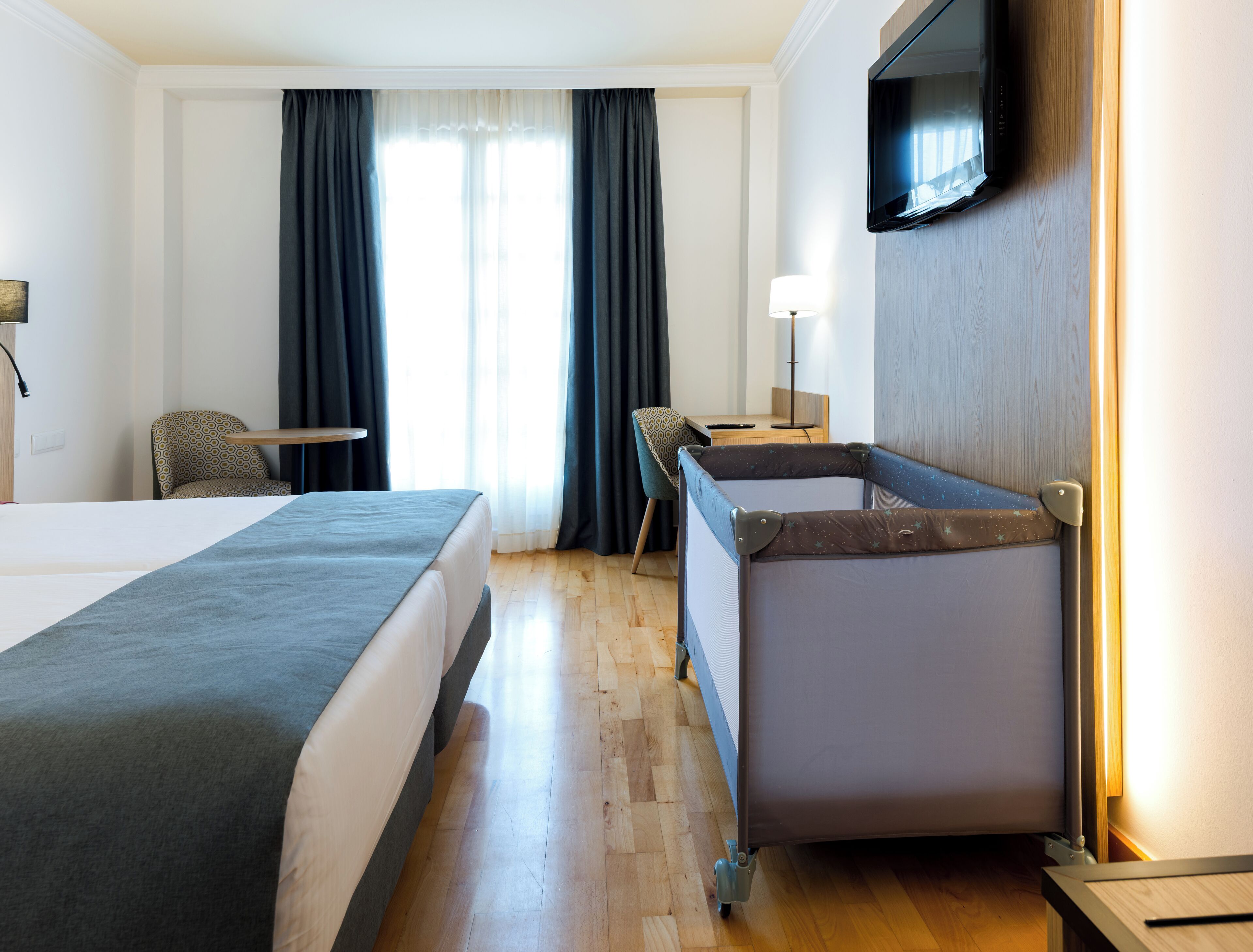 A hotel room with a large bed, a desk with a lamp, a TV mounted on the wall, and a small seating area with a chair.