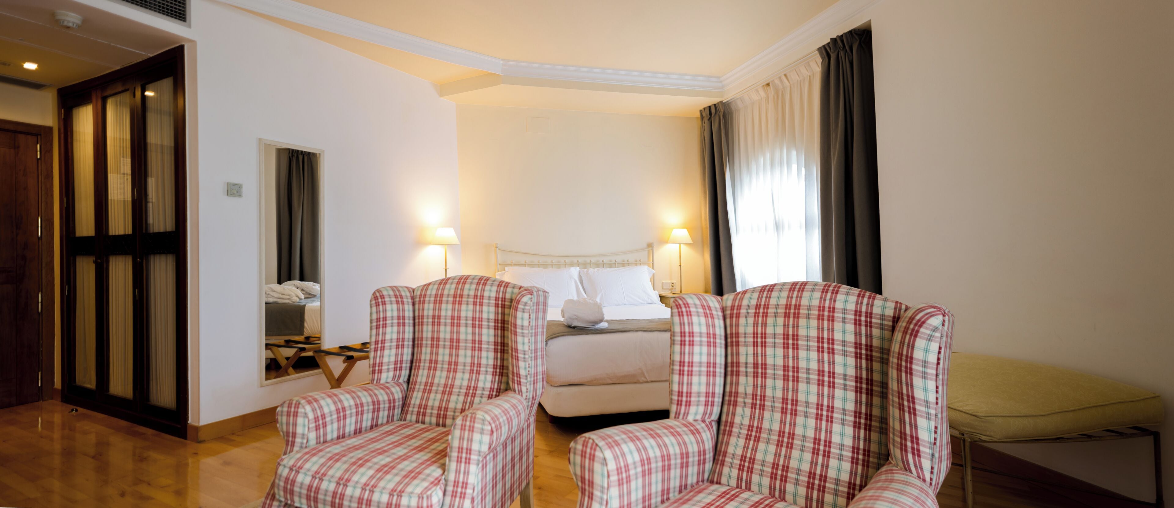 Una habitación de hotel con una cama, dos sillones a cuadros, una mesita de noche y un espejo.