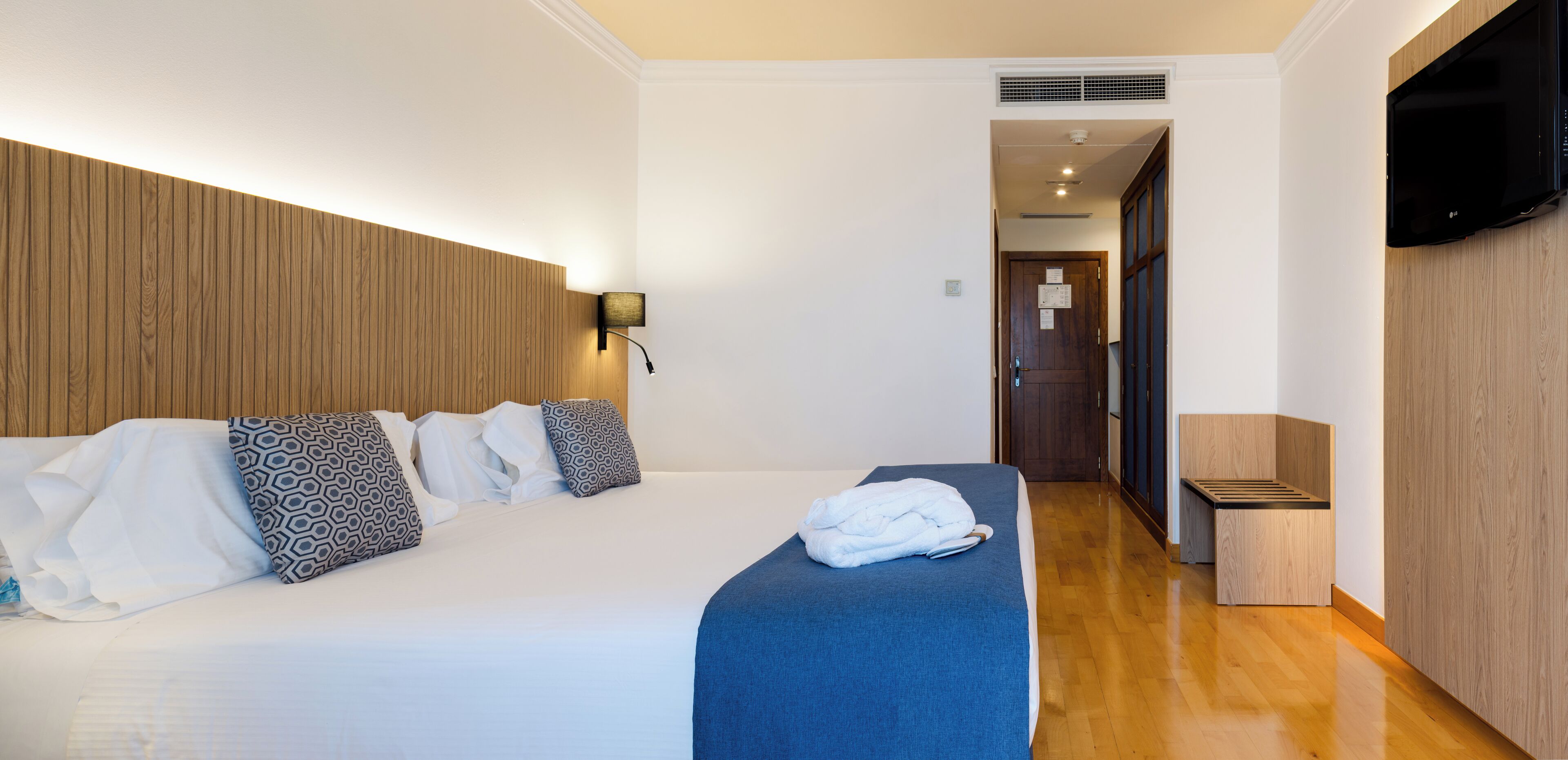 A hotel room with a bed, a TV mounted on the wall, and a wooden headboard.