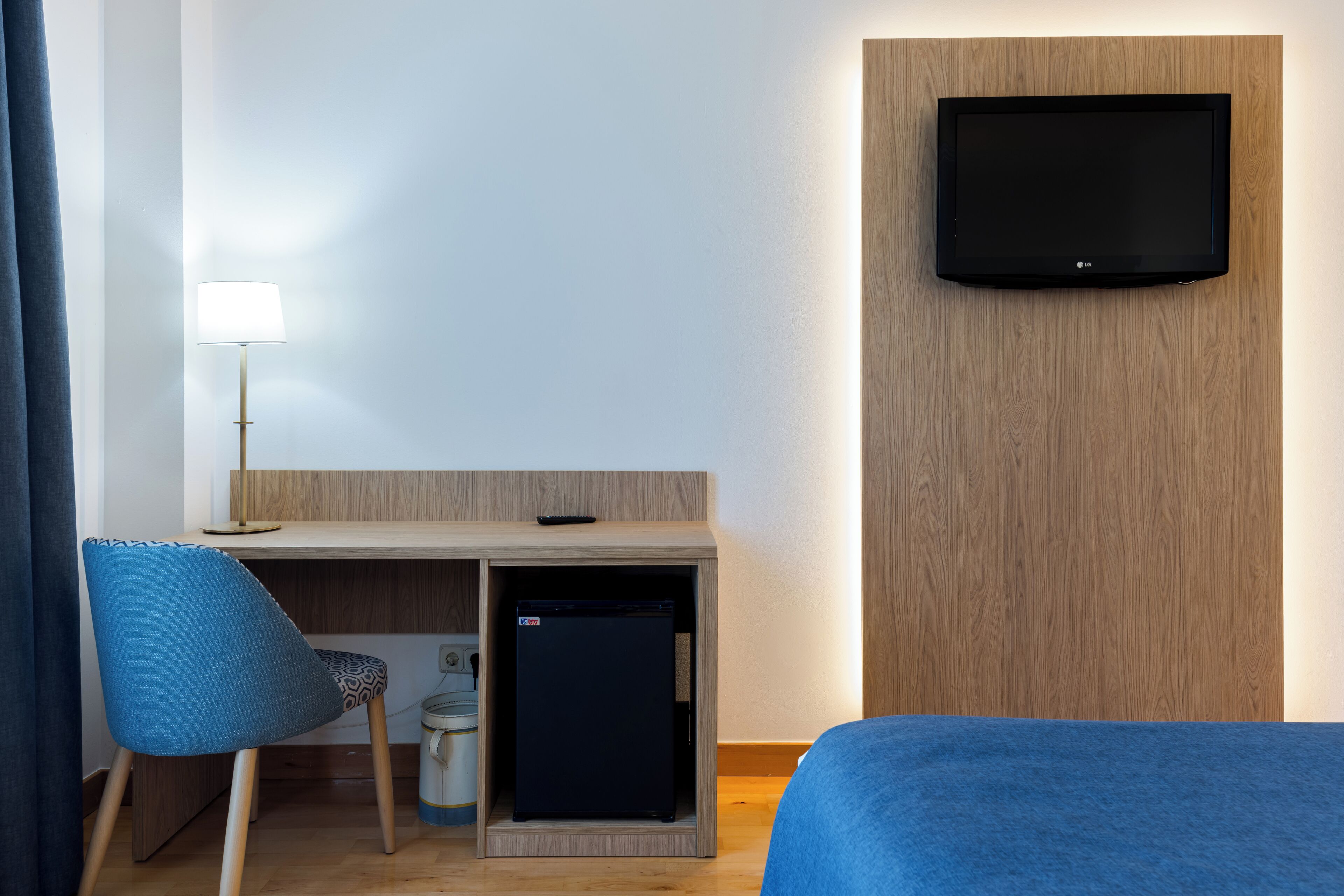 Habitación de hotel con escritorio, una silla, televisor montado en la pared y un refrigerador pequeño.