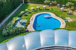 Una piscina de diseño curvo, rodeada de sillas de descanso y sombrillas, con una estructura de invernadero al frente.