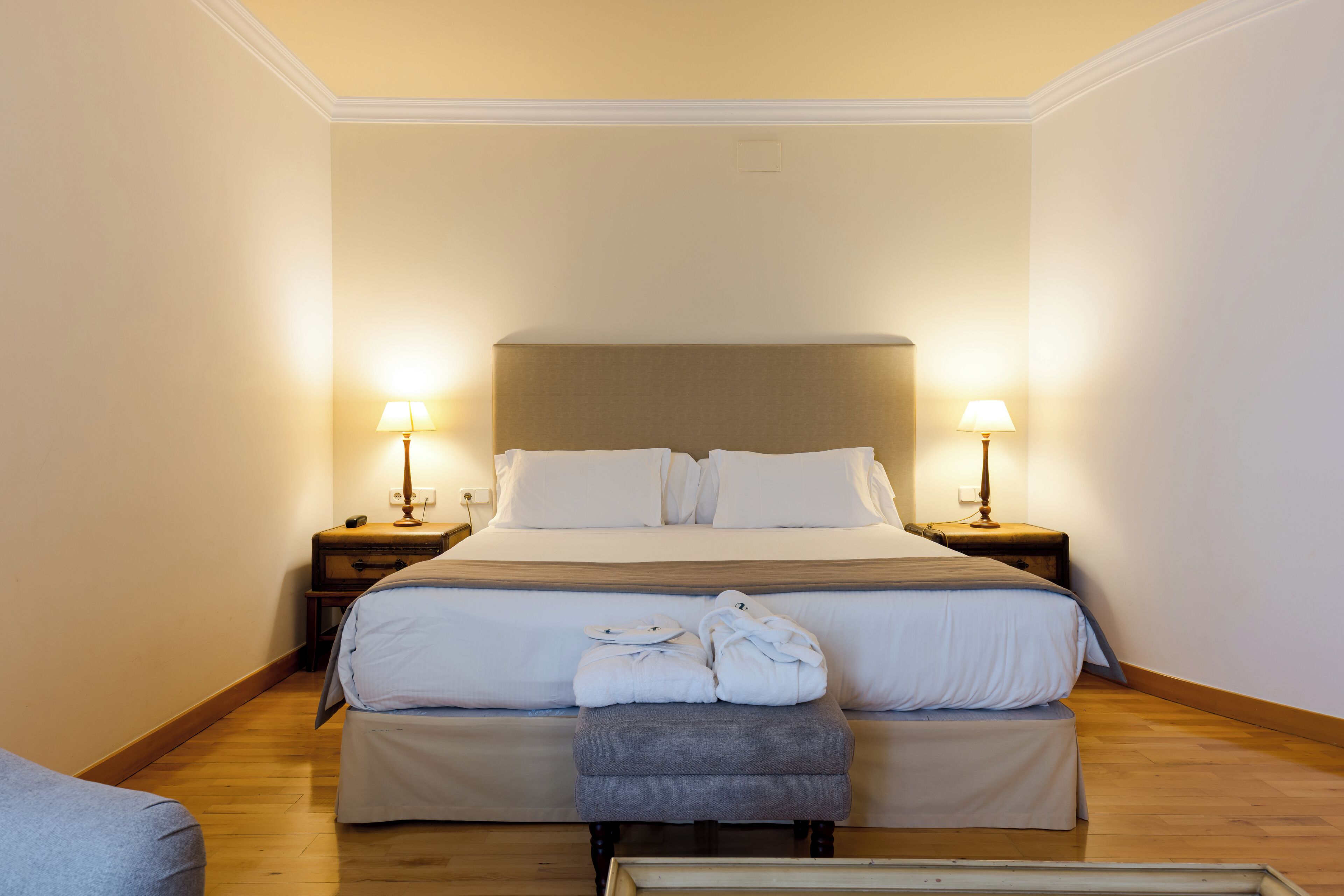 Habitación de hotel con una cama grande, dos mesitas de noche con lámparas, un banco y una pequeña mesa con lámpara.