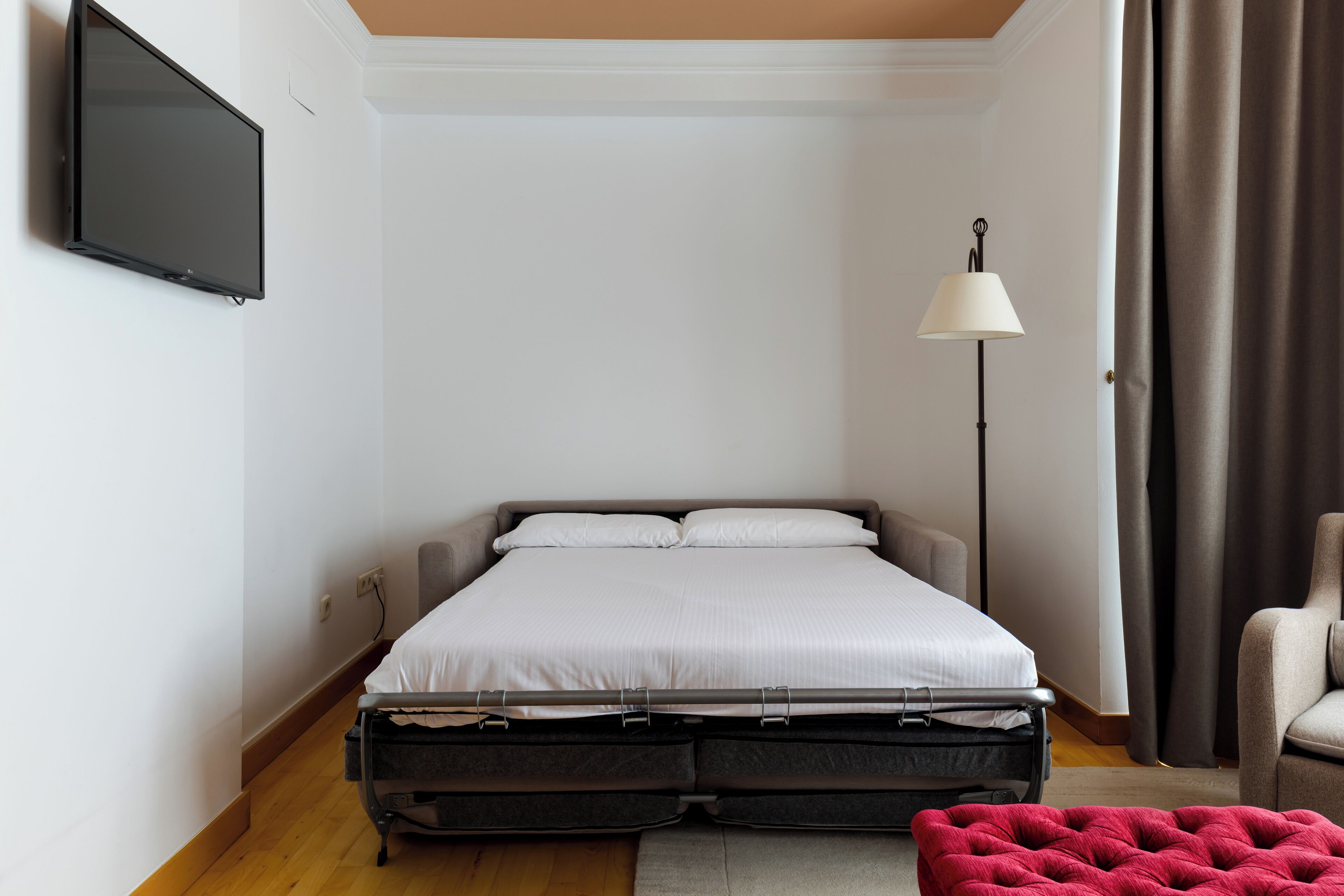 Un dormitorio ordenado con una cama, un televisor de pantalla plana montado en la pared, una lámpara de pie y una silla.