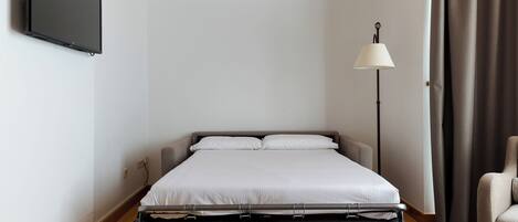 Un dormitorio ordenado con una cama, un televisor de pantalla plana montado en la pared, una lámpara de pie y una silla.