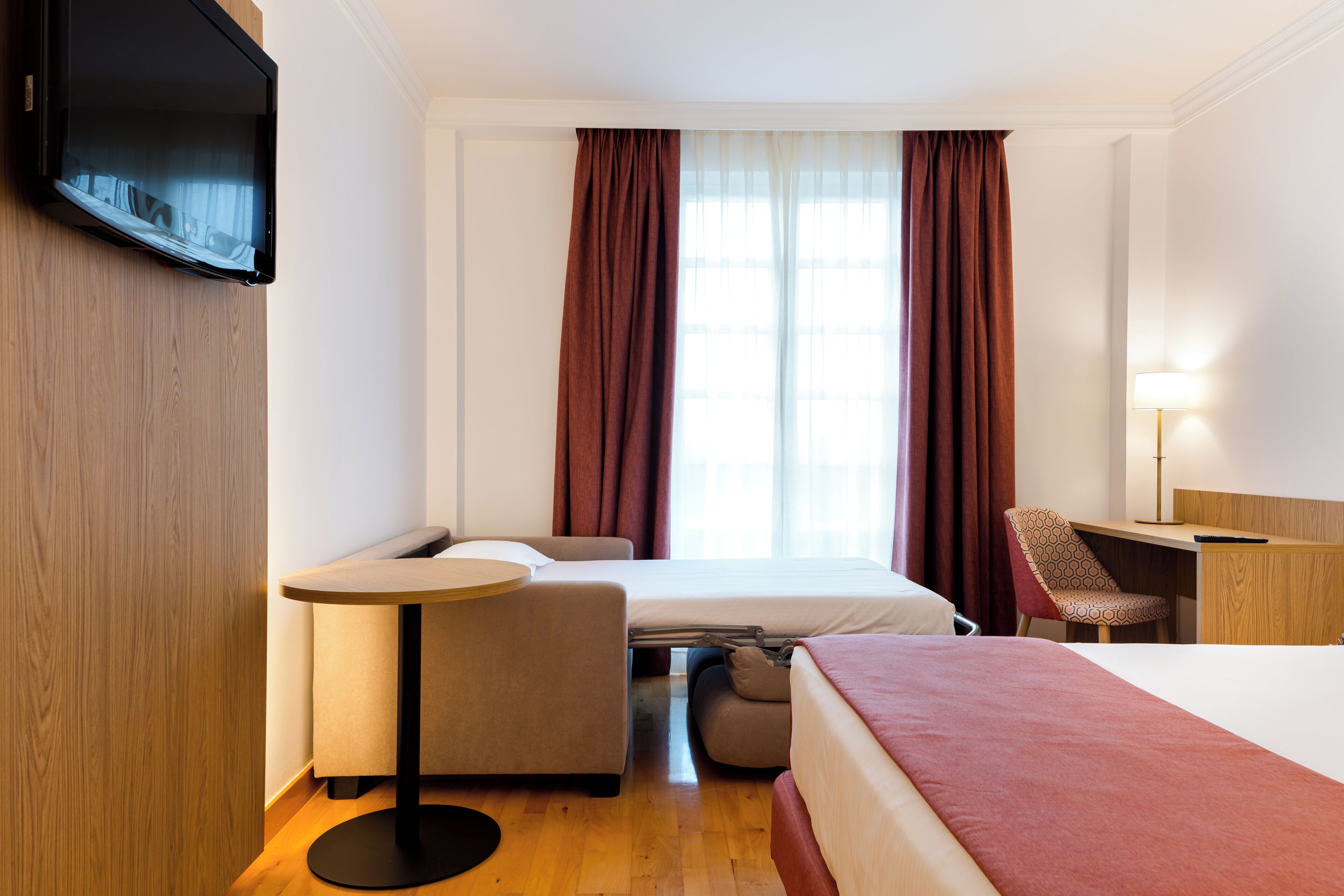 A hotel room with a bed, a desk, a chair, a small table, and a TV mounted on the wall.