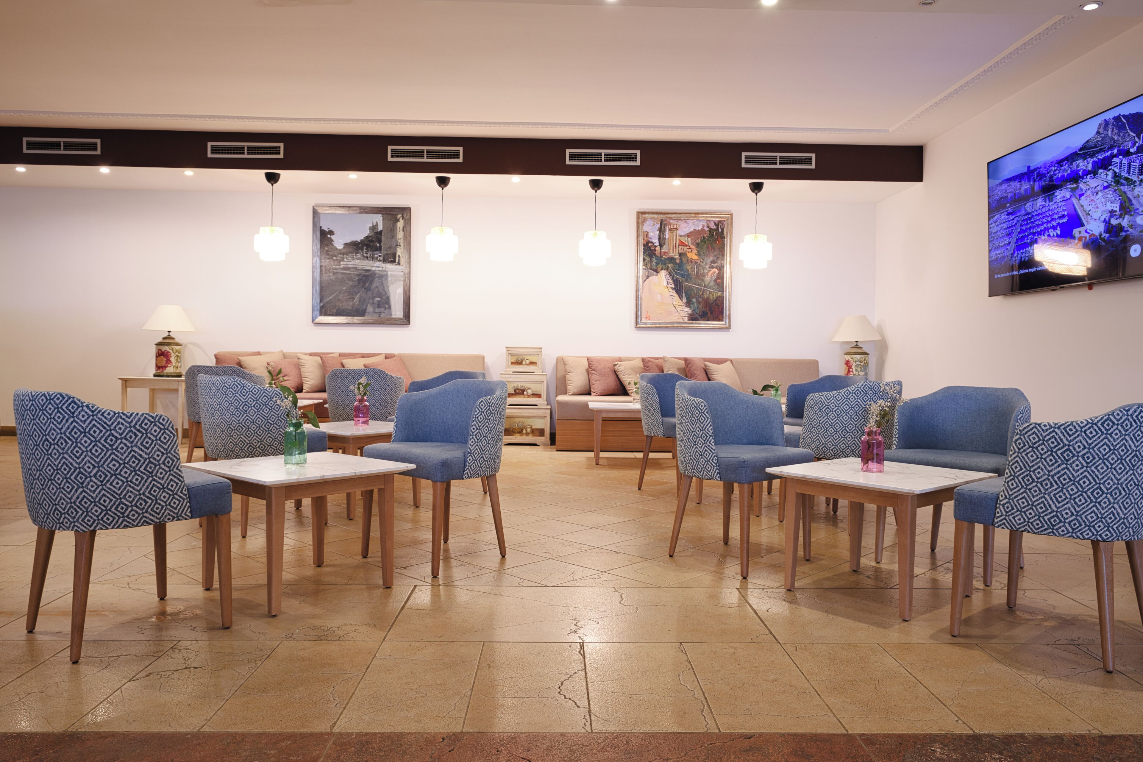 A modern hotel lobby with blue patterned sofas, wooden tables, and a large TV screen on the wall.