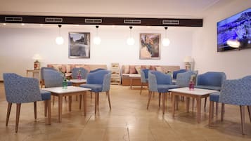 A modern hotel lobby with blue patterned sofas, wooden tables, and a large TV screen on the wall.