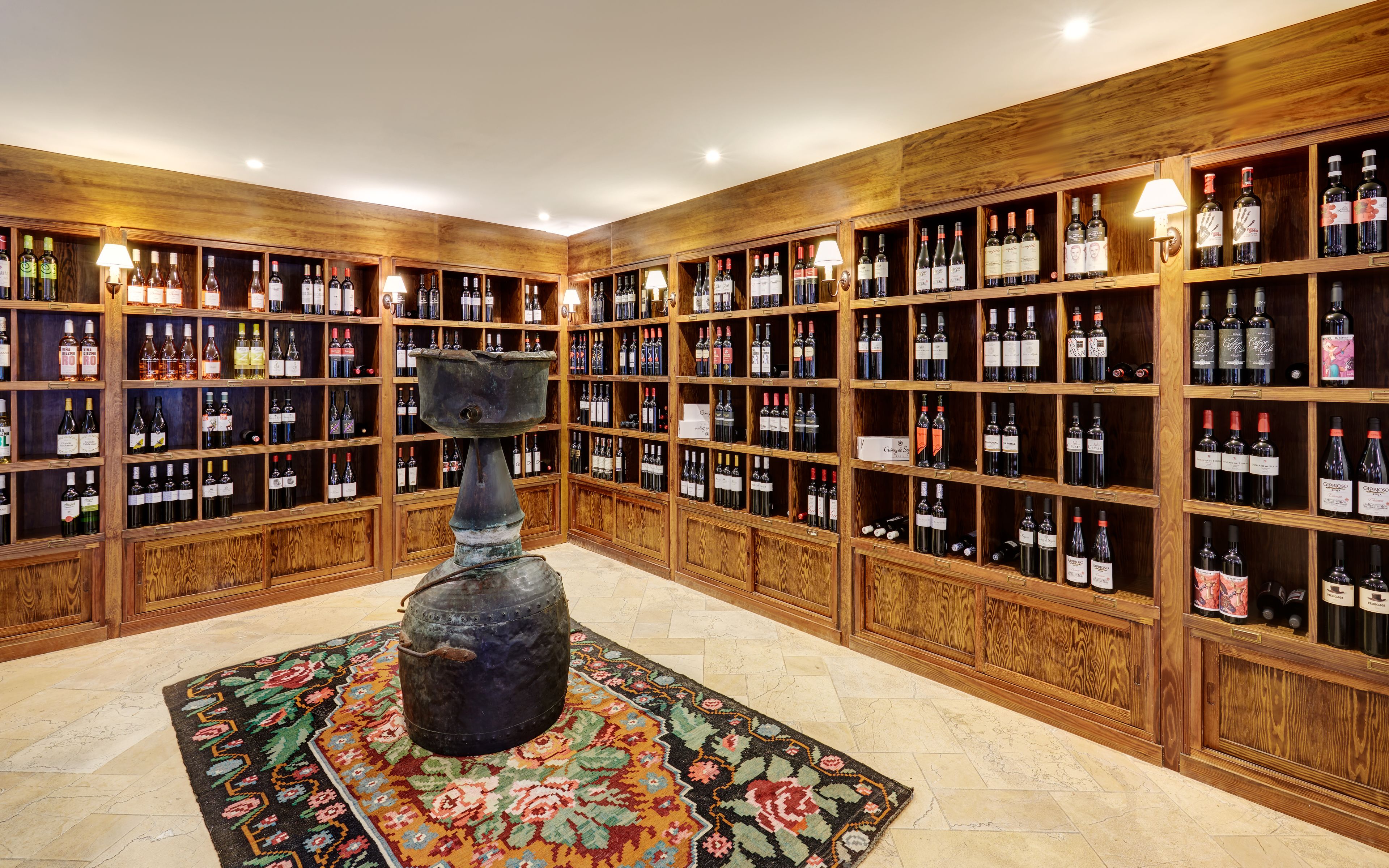 A wine cellar with wooden shelves filled with bottles, a central decorative urn, and a patterned rug on the floor.