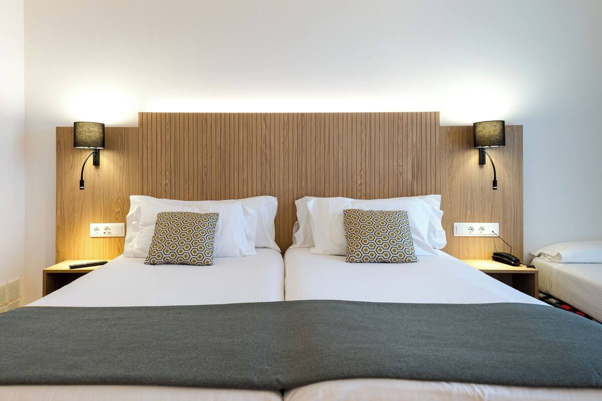 Un lit double avec une tête de lit en bois, deux tables de chevet avec des lampes et un lit simple sur la droite.