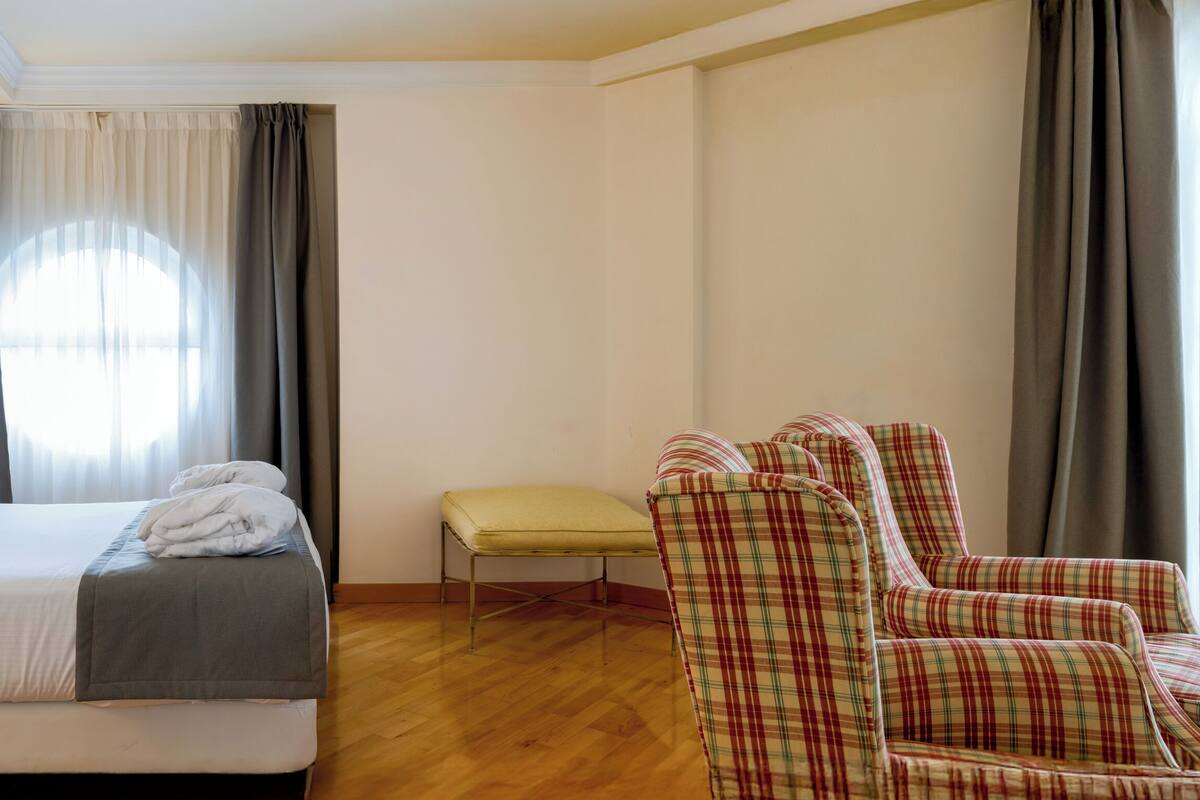 Une chambre d’hôtel avec un lit, des lampes de chevet, une chaise, un banc et une fenêtre avec des rideaux.