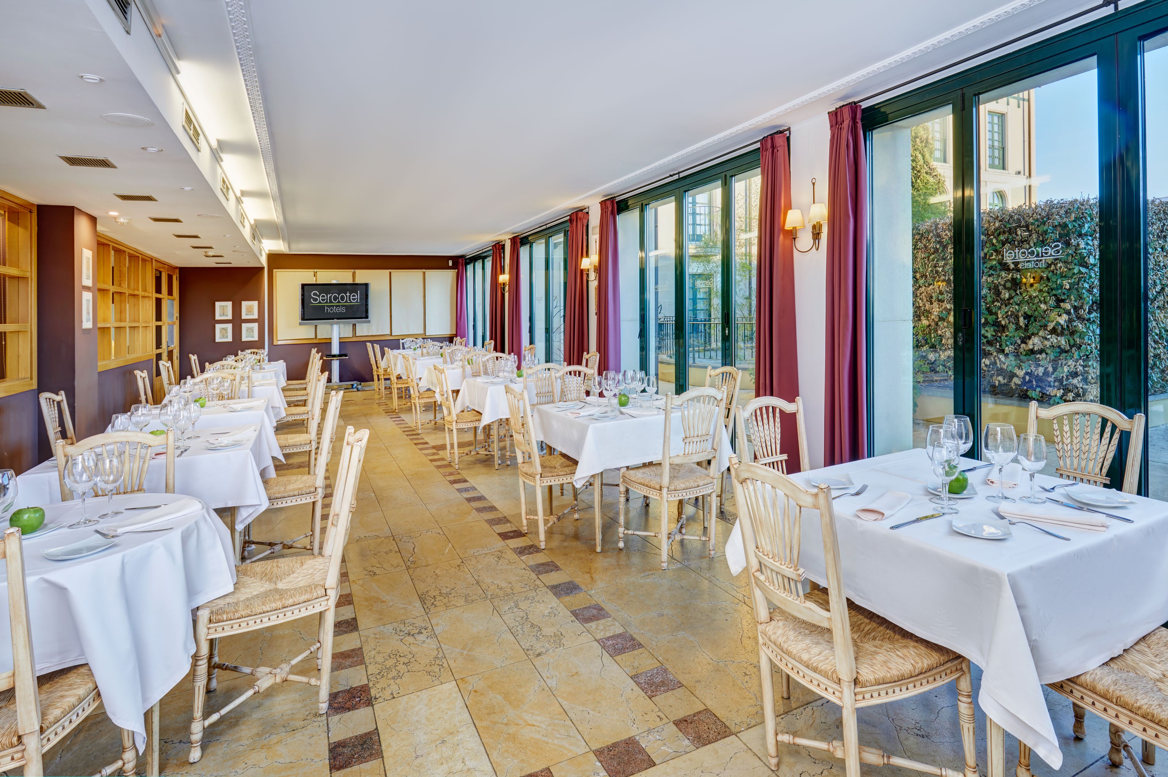 A long dining room with multiple tables set for dining, large windows, and a sign for 'Sercotel hotels'.