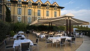 Un área de comedor al aire libre con mesas y sillas, un paraguas grande y un edificio con ventanas arqueadas al fondo.