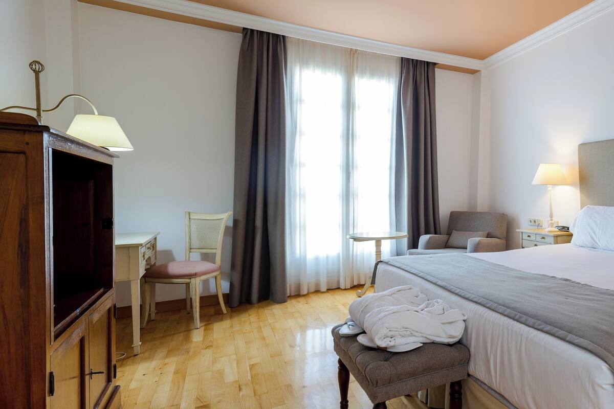 Une chambre d’hôtel avec un grand lit, des lampes de chevet, une armoire en bois, une chaise et un banc.