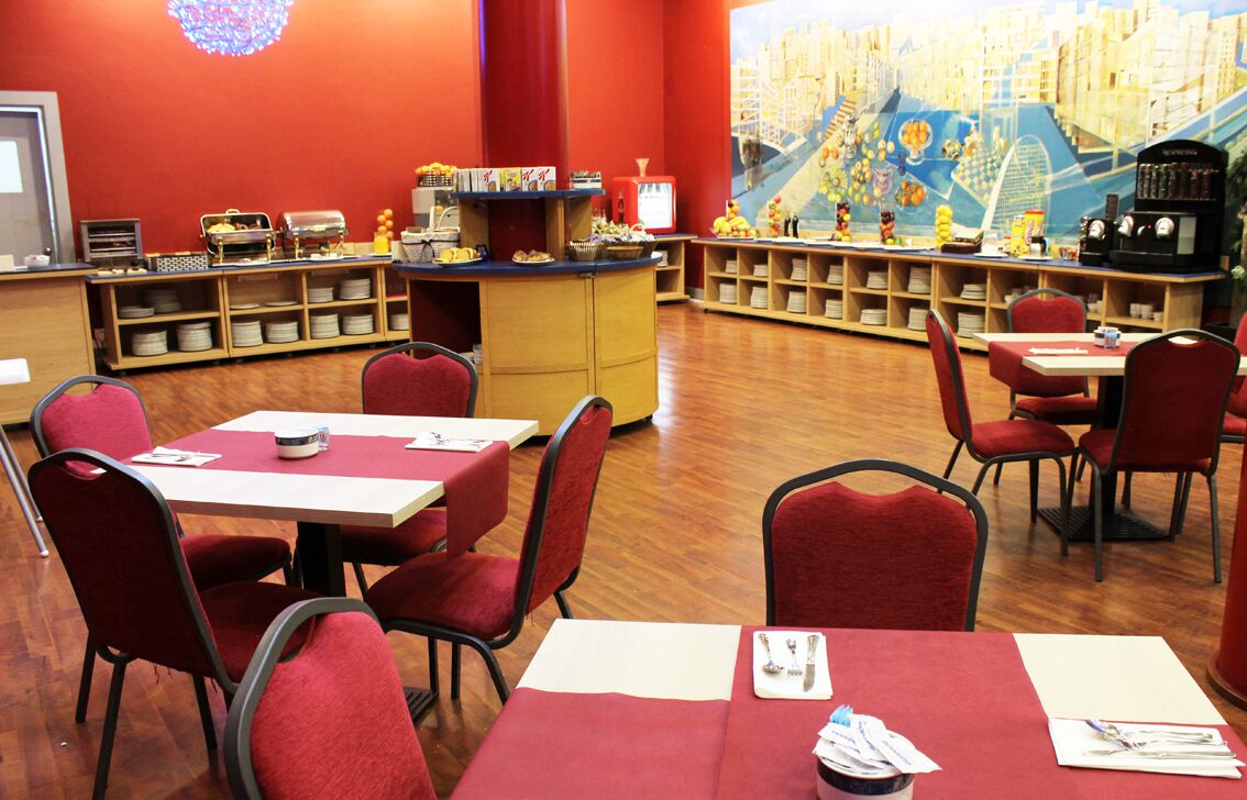 Un restaurante con sillas y mesas rojas, un área de bufé con comida y bebidas, y un mural en la pared.