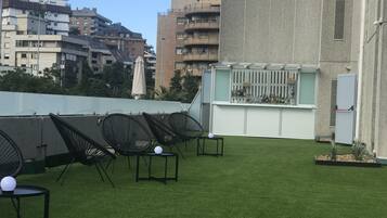Terraza en la azotea con césped artificial, sillas de metal negro y una zona de bar con botellas.