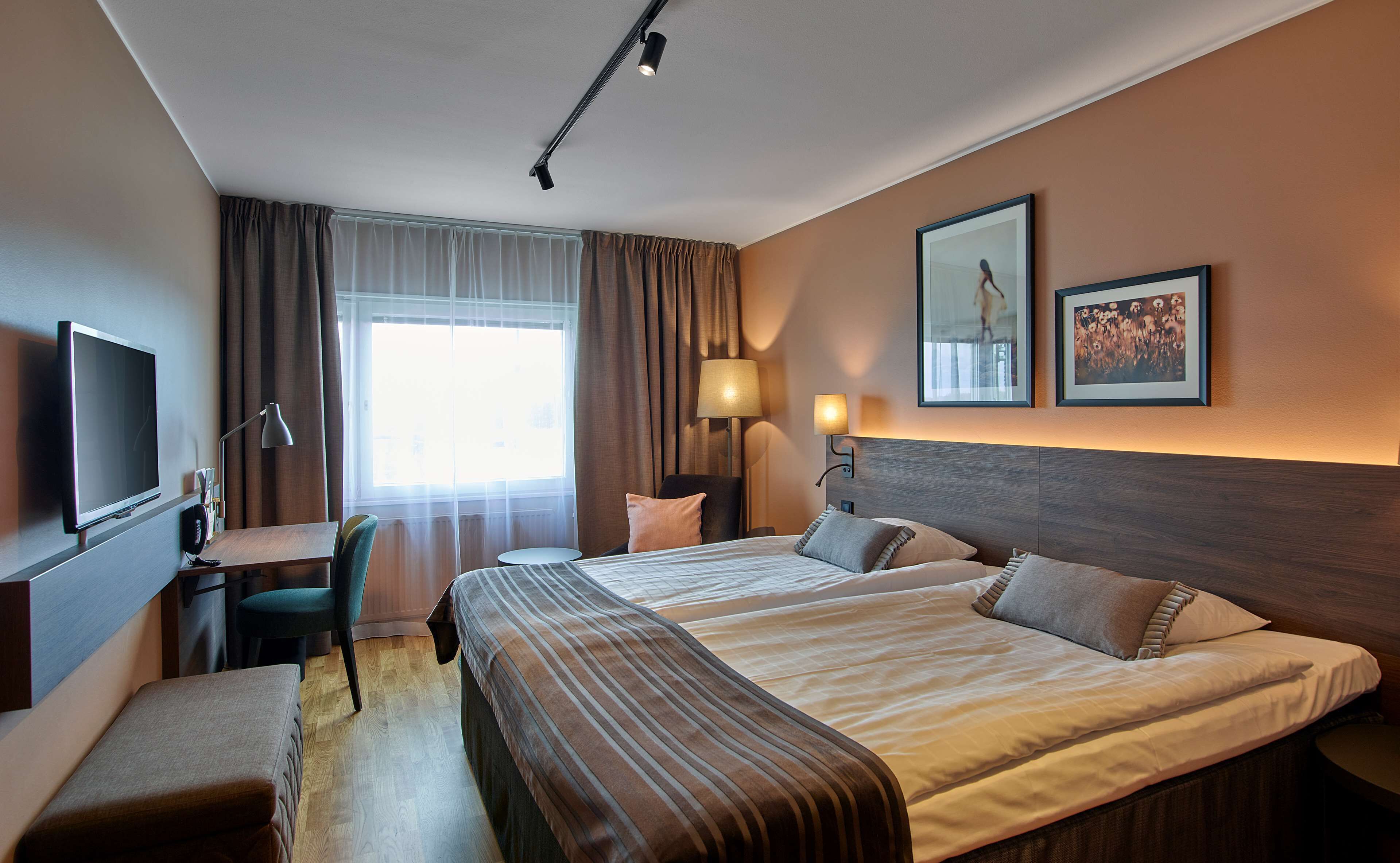 Une chambre d’hôtel avec un grand lit, un bureau avec une chaise, une télévision et deux tableaux encadrés au mur.